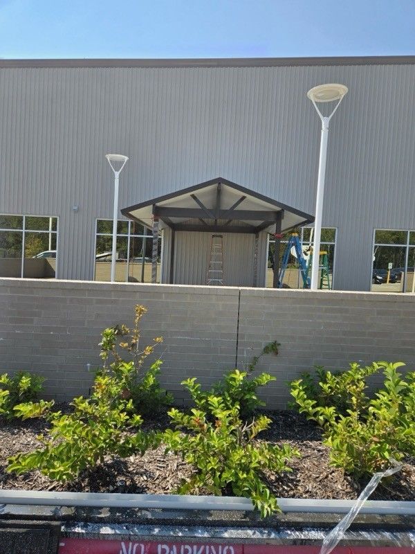 Exterior view of a building with a canopy entrance, two tall street lamps, and a low brick wall with greenery.