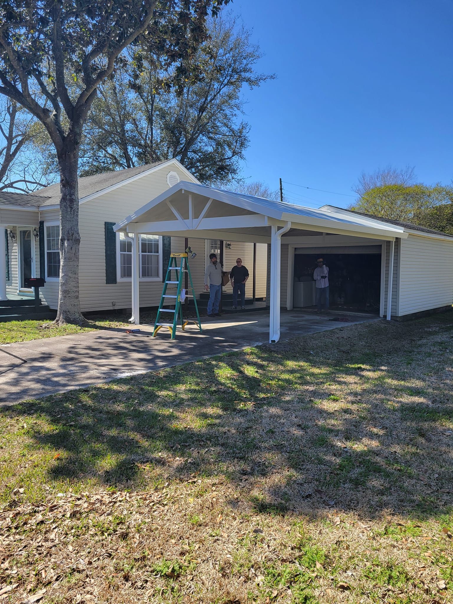 House with carport; people stand under carport, ladder leans against a post. Sunny day with brown grass and blue sky.
