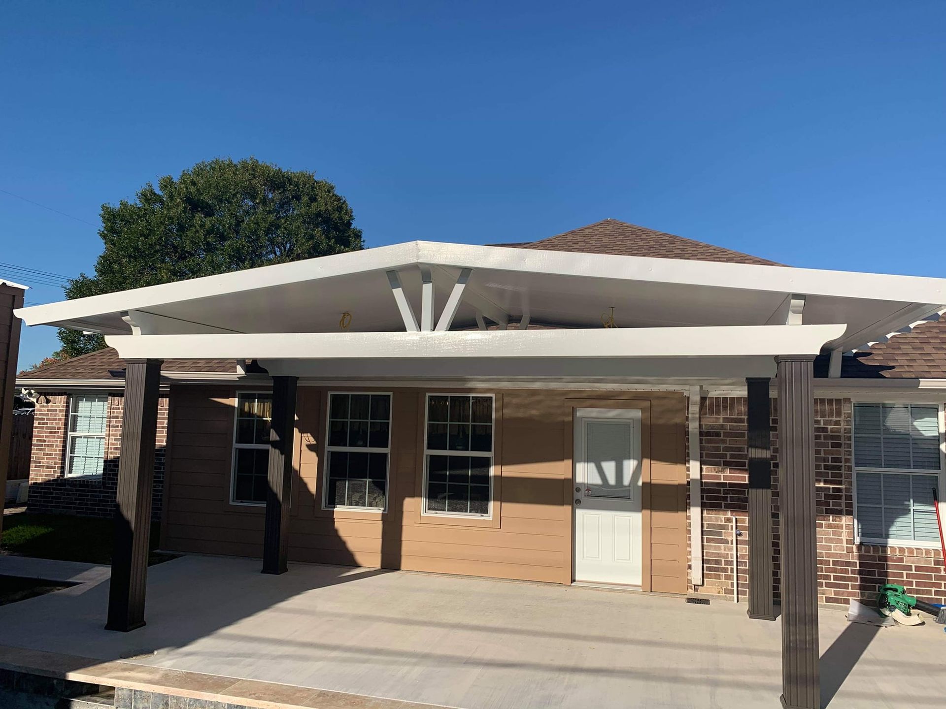 Covered patio with white roof and dark brown supports, attached to a brick building with windows and a door under a blue sky.
