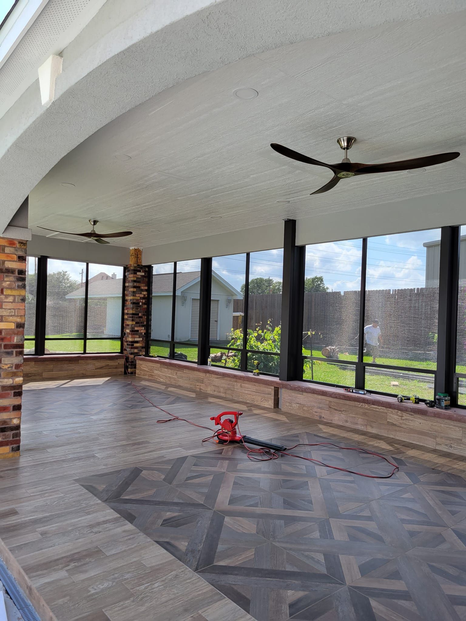 A screened-in patio with a patterned floor, ceiling fans, and a brick accent.  The patio overlooks a grassy yard and residential buildings.