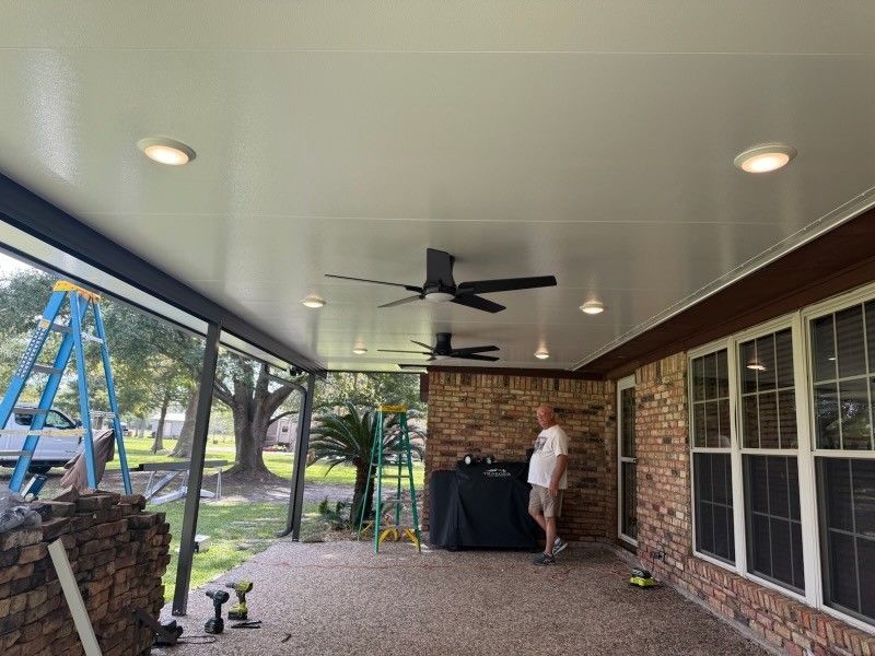 A covered outdoor patio with a man, fans, recessed lights, and a brick wall.