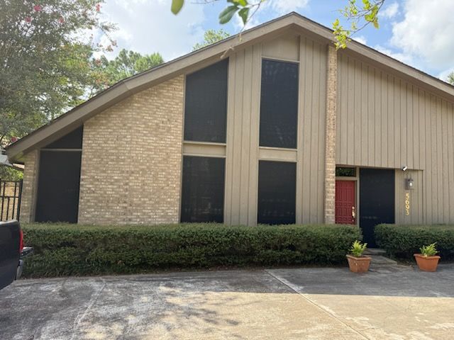 A light-colored brick and beige-sided building with black-screened windows and a red door. A well-trimmed hedge lines the front.