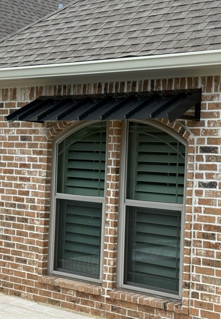 Black awning above a tall arched window with closed green shutters, set in a brick wall.
