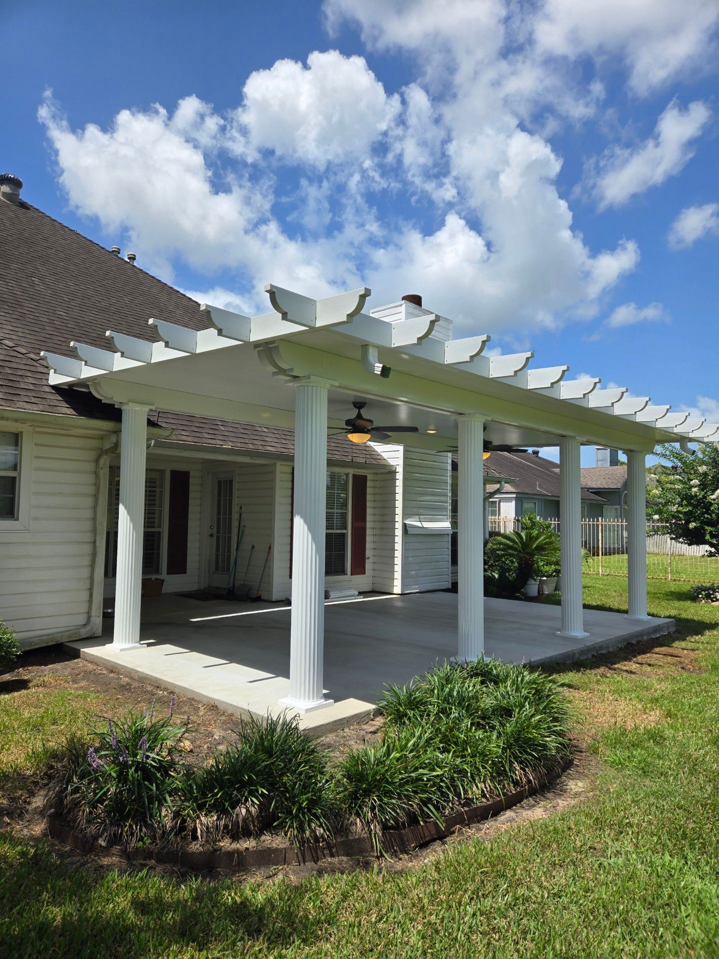 White pergola attached to a house with a concrete patio. Lush green grass and bushes surround the patio on a sunny day with clouds.