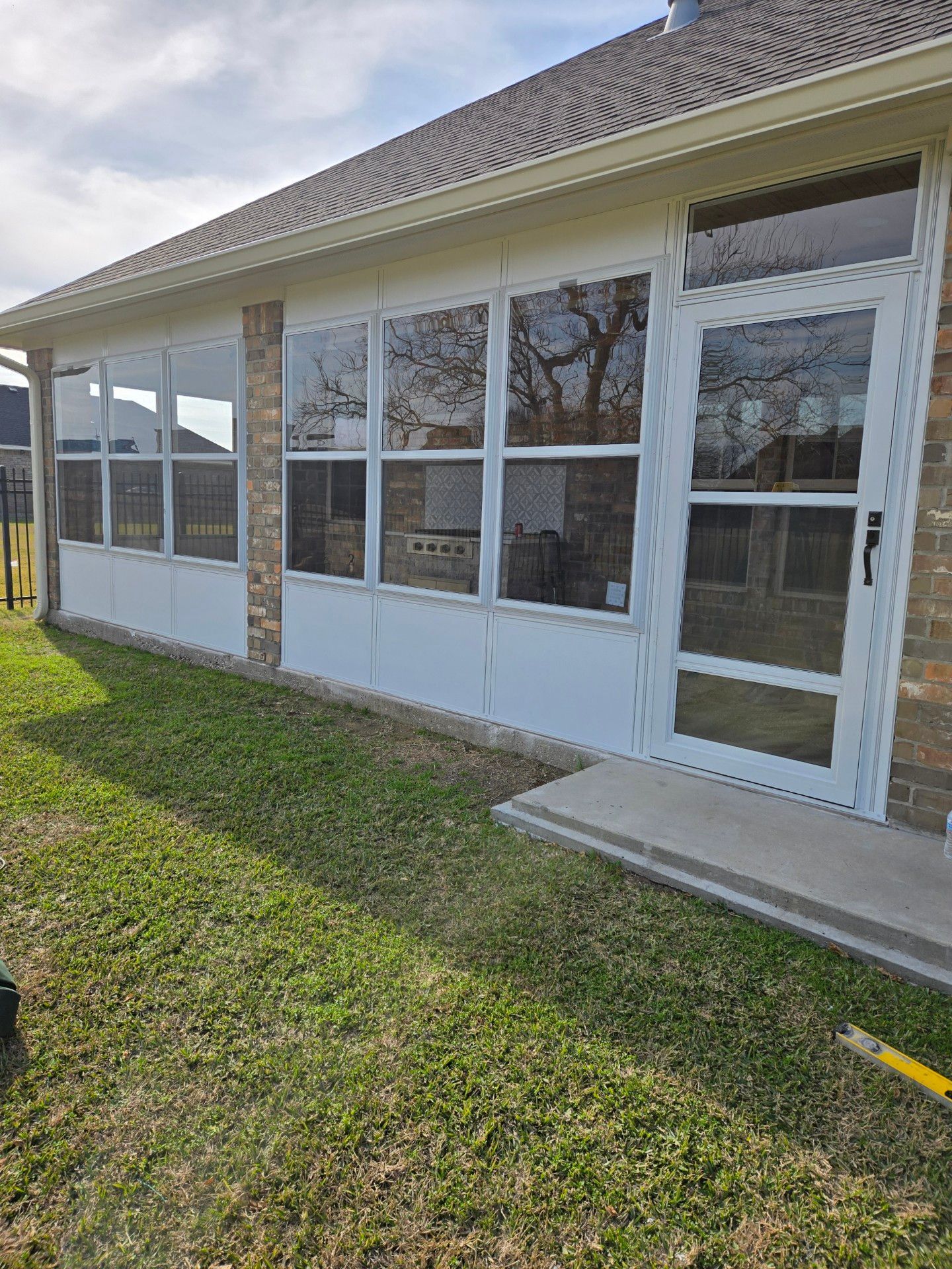 A sunroom addition to a brick house with white framed windows and door. Green grass surrounds the room.