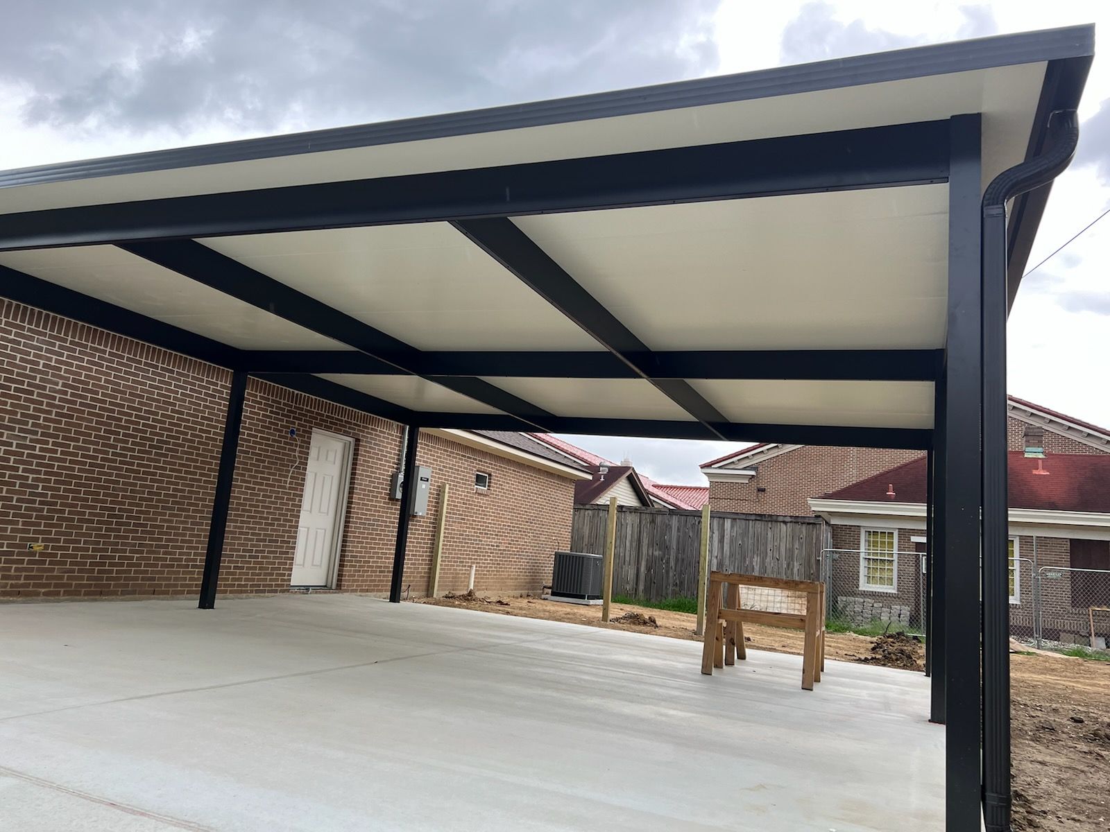 A newly constructed carport with a black metal frame, white ceiling, and concrete floor; houses and a fence are visible in the background.