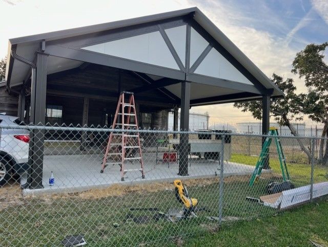 A partially constructed metal pavilion with a concrete base and a red ladder sits on a sunny day.