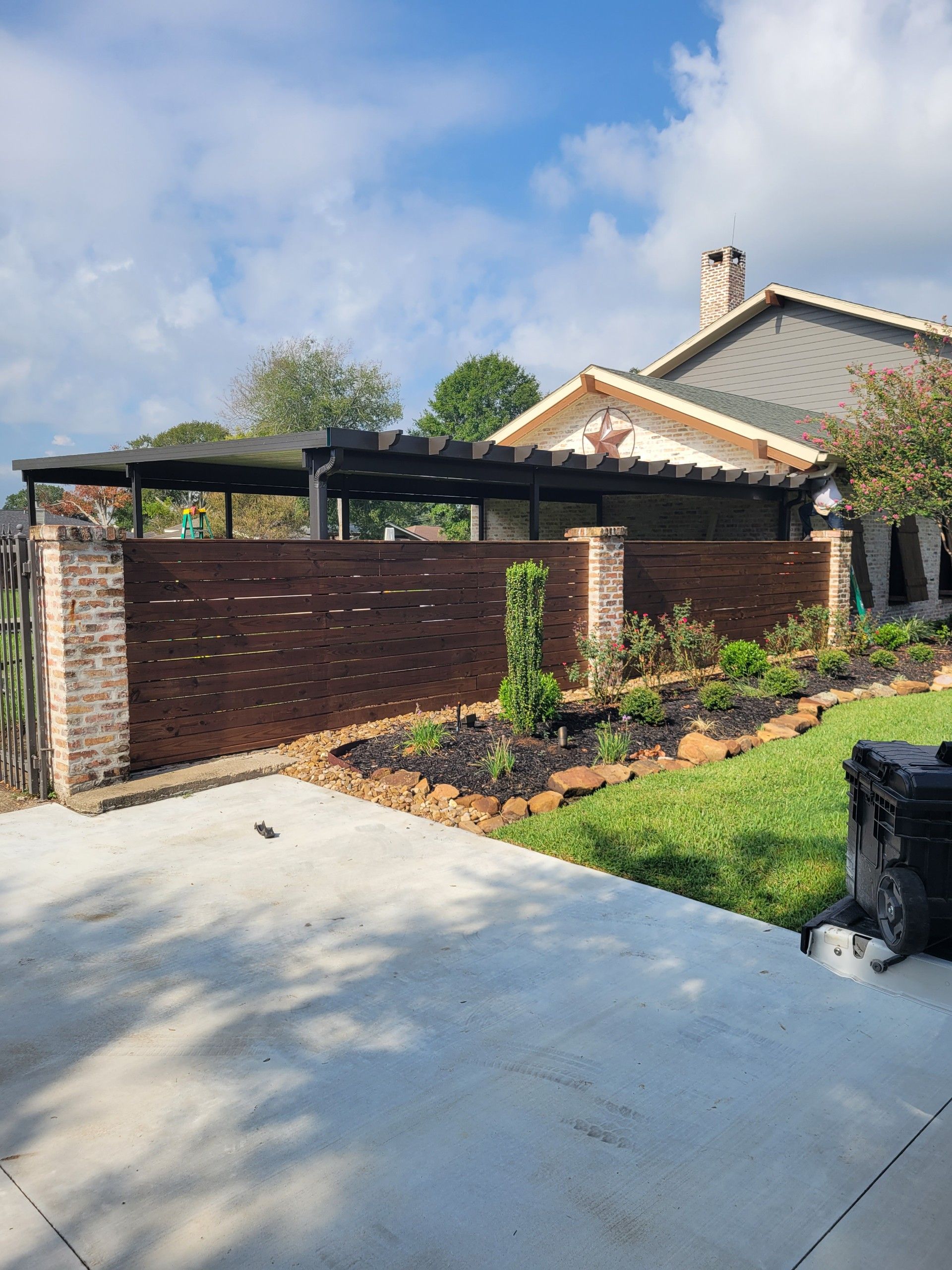 A modern dark wooden fence with brick pillars, in front of a house with a concrete driveway and green lawn under a blue sky.