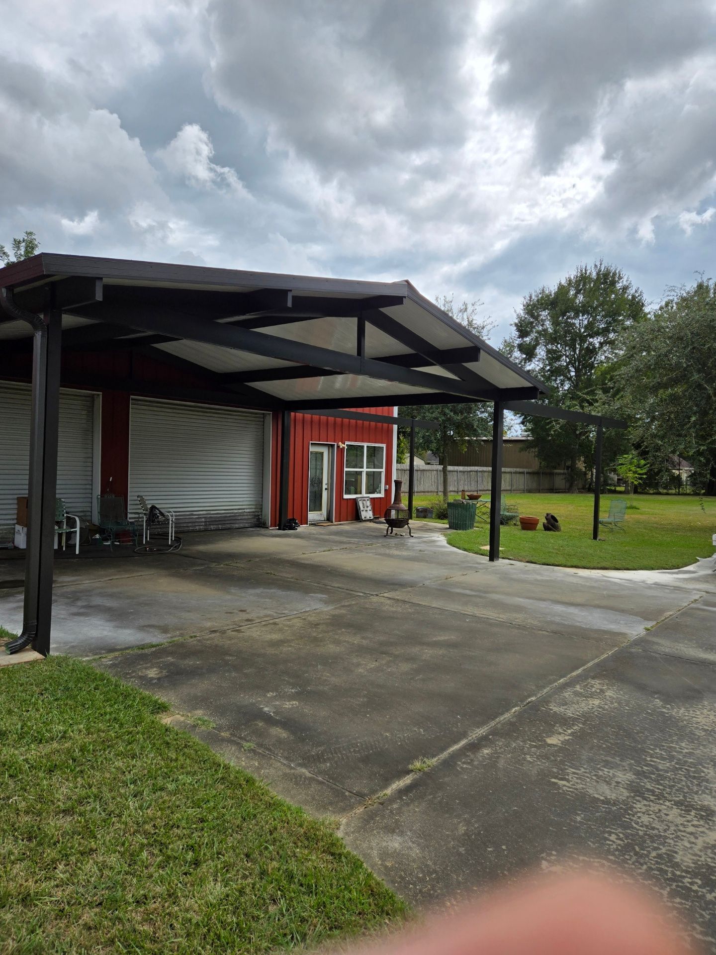 Red building with a black carport on a concrete driveway under a cloudy sky; green grass borders the driveway.