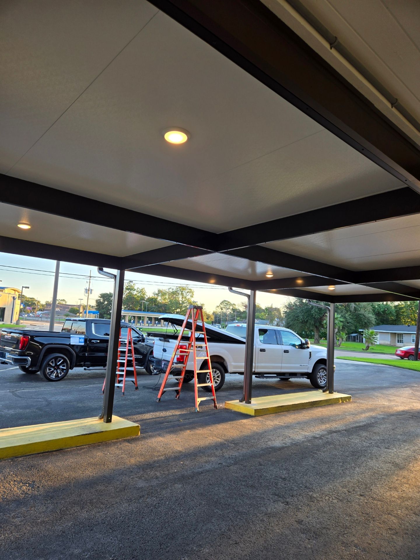 Carport with black and white roof, yellow curbs, and parked trucks. A ladder is in use near a pickup truck, possibly working on lights.