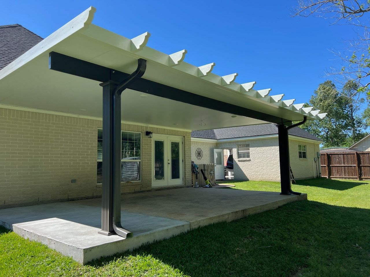 A covered patio with a concrete floor and dark columns attached to a light brick house on a sunny day.
