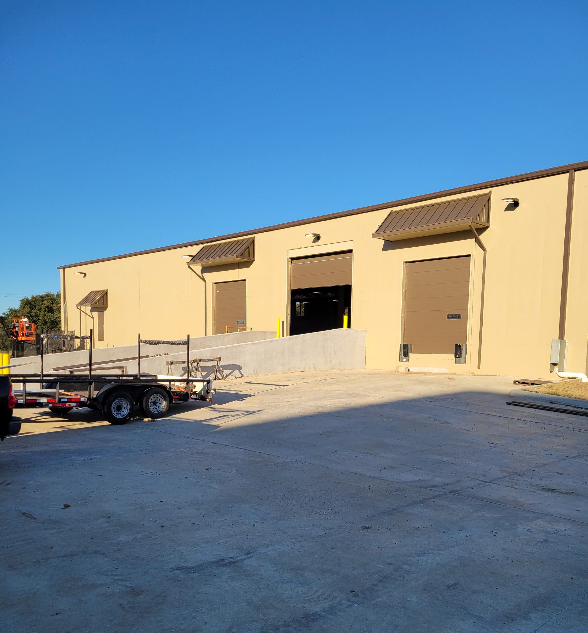 Tan industrial building with three loading doors, a trailer in front, under a clear blue sky.