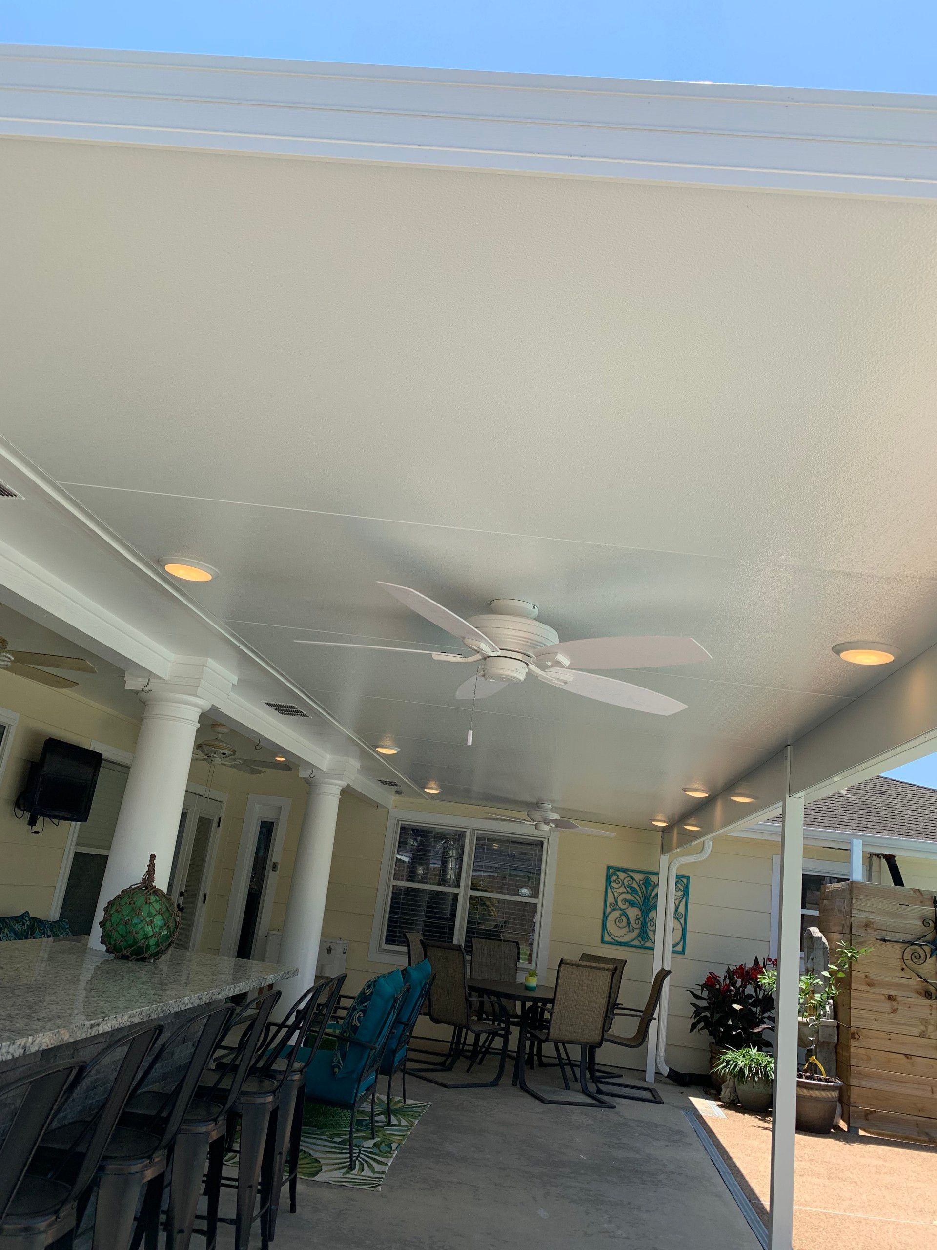 An outdoor patio with a white ceiling, ceiling fan, and bar.  Tables and chairs are set up beneath the covered area.