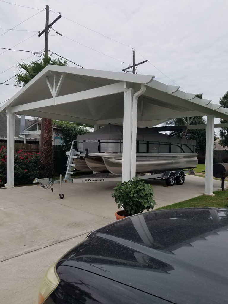 White carport shelters a pontoon boat on a trailer, on a concrete driveway. Gray sky.