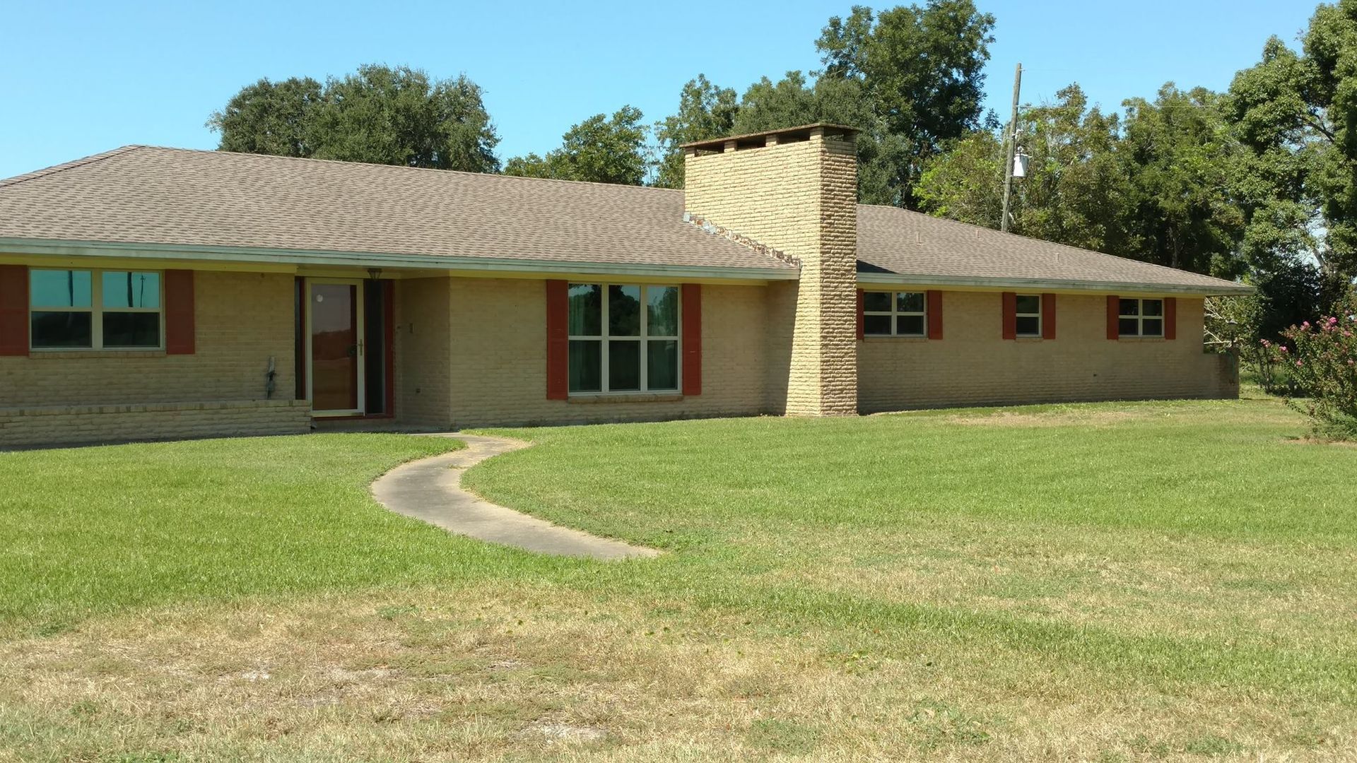 A single-story brick house with a chimney, a winding sidewalk, and red shutters. The house sits on a grassy lawn with trees in the background.