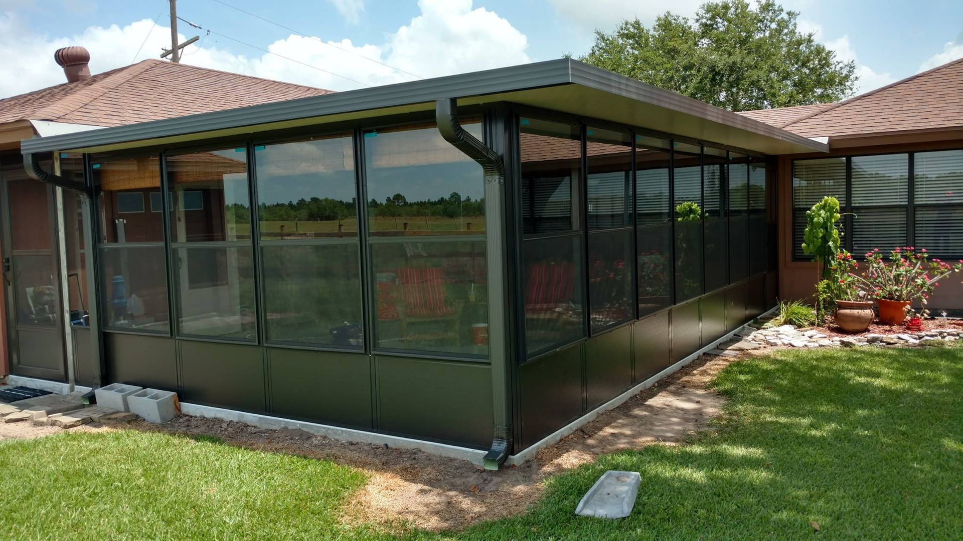Sunroom attached to a house with black frames and glass windows. It has a green lawn and a cloudy sky.