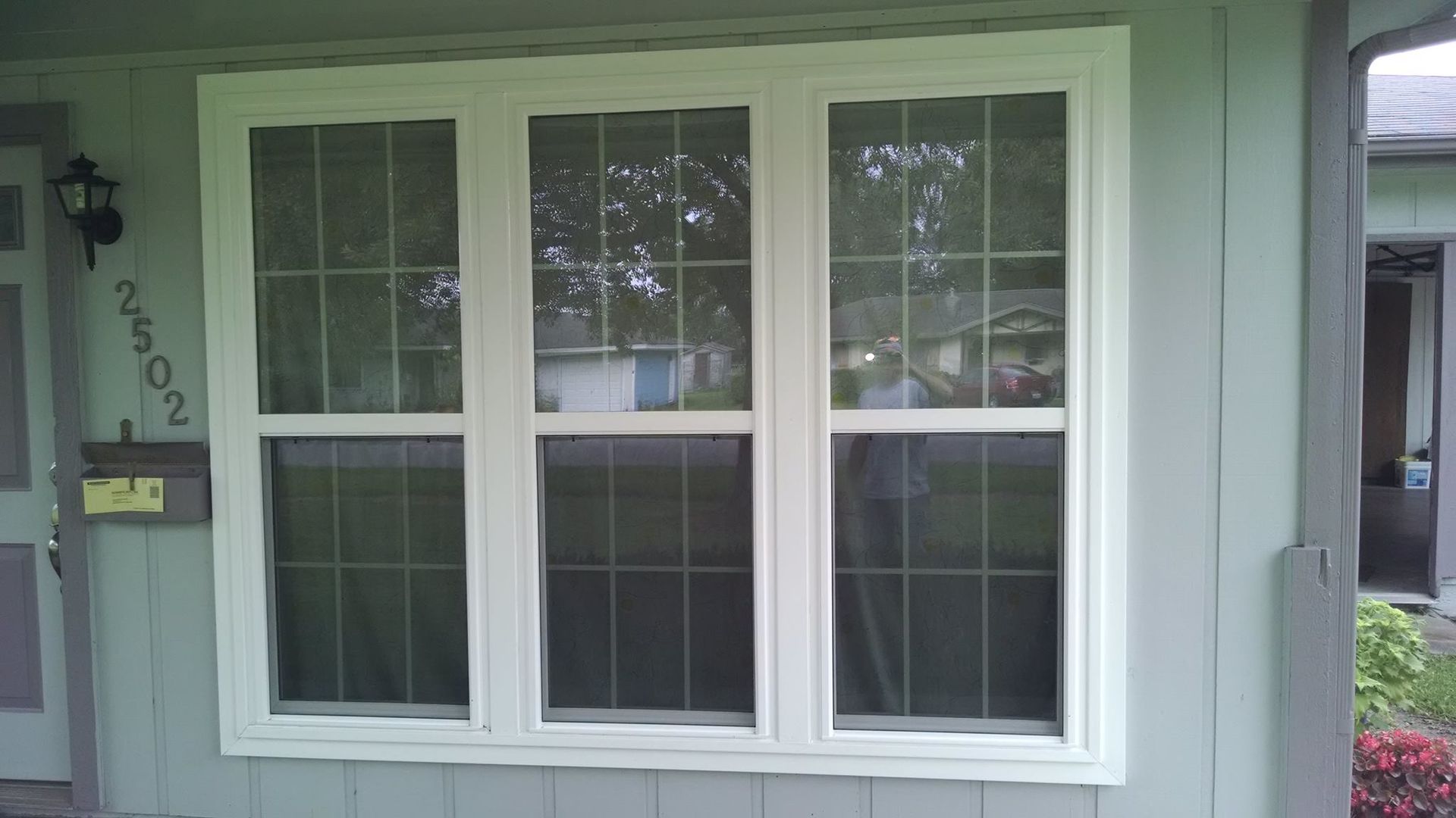 Triple-pane white-framed window with gridded panes on a light-green house, with a black light fixture and the number 