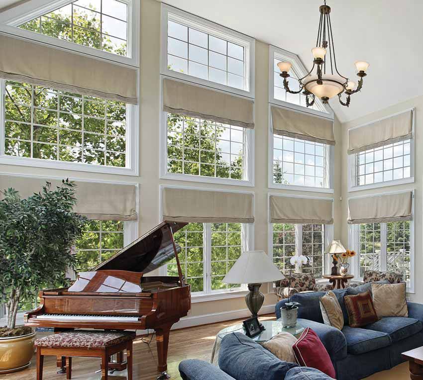 Living room with tall windows, piano, and blue sofas; sunlight streams in. Beige blinds and a chandelier add to the bright space.