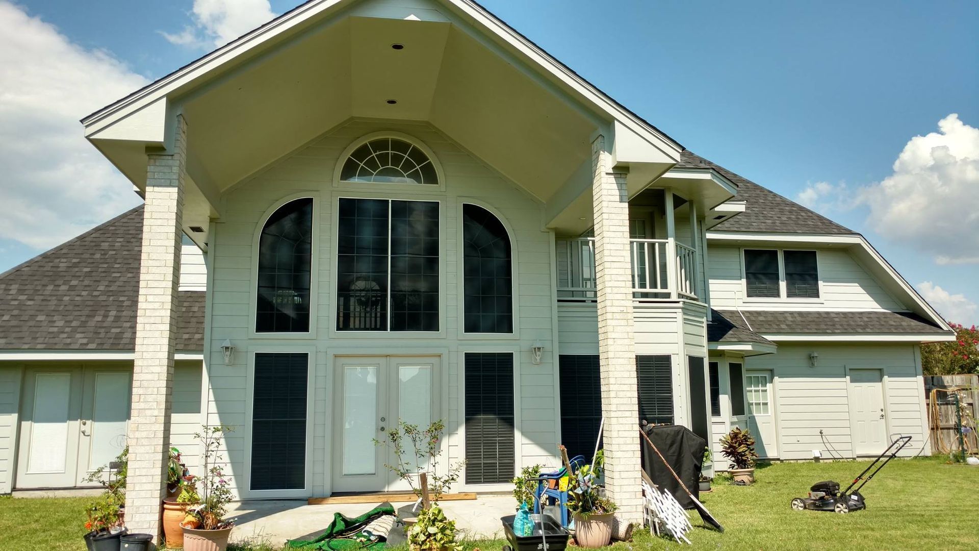 White two-story house with large front windows and a gabled roof. Green lawn, blue sky, and gardening tools are in front.