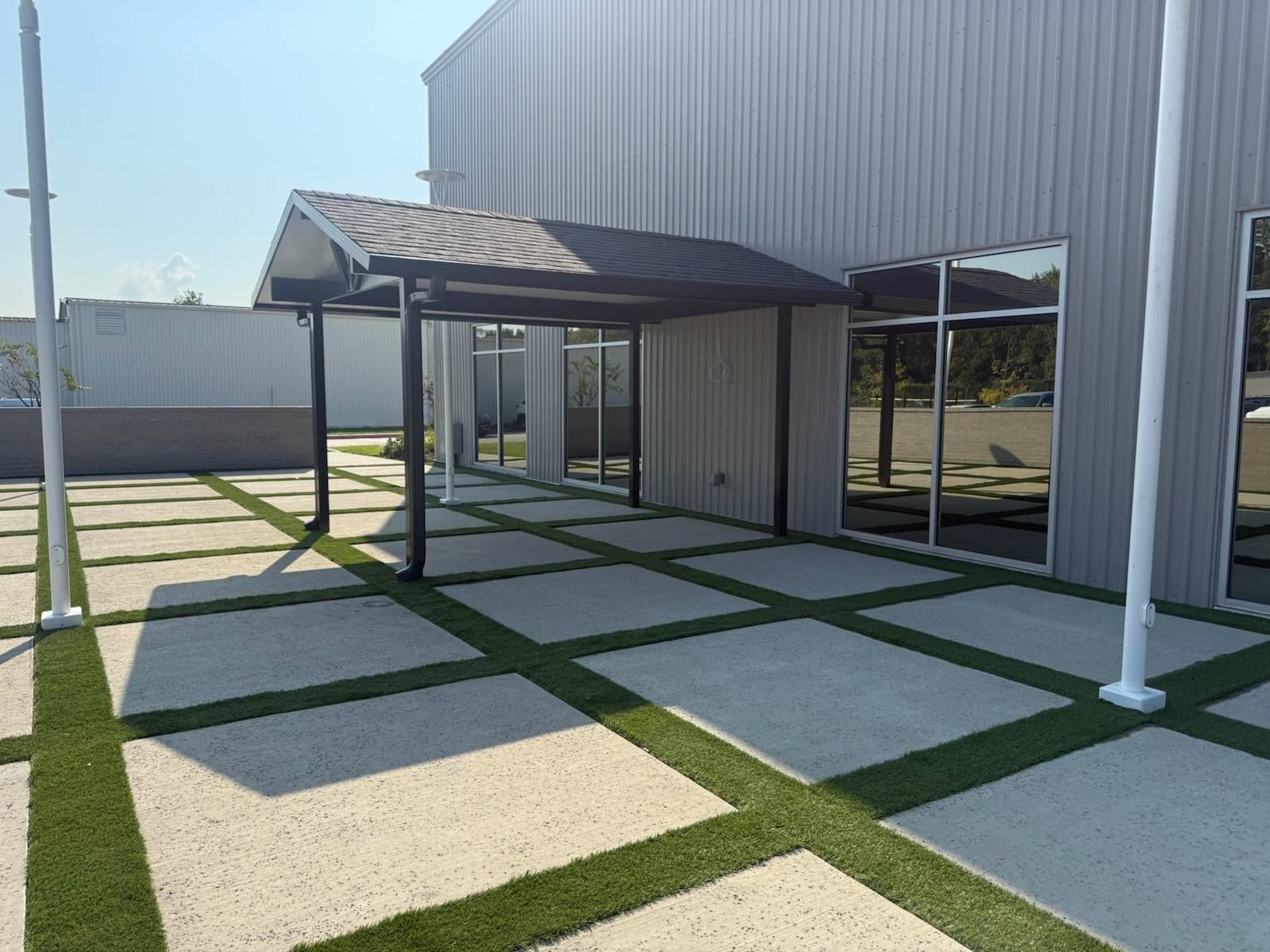 Exterior of a building with a walkway of concrete squares separated by artificial turf; a covered entrance.