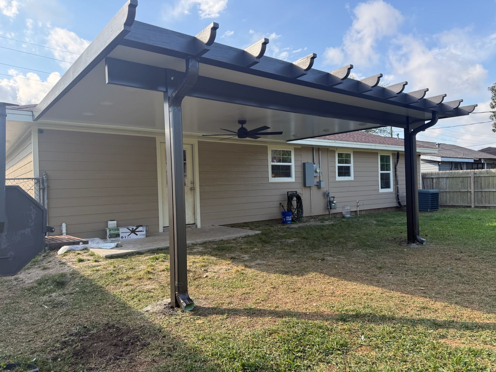 Backyard patio with a dark pergola, attached to a tan house. Green grass and blue sky.