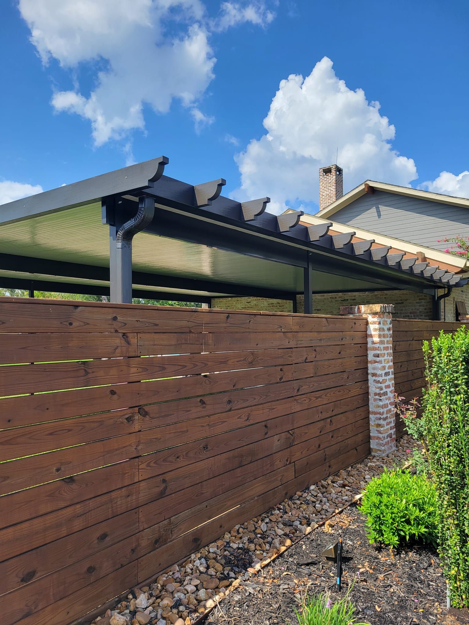 A dark wooden fence with a pergola and brick chimney in front of a blue sky with fluffy clouds.