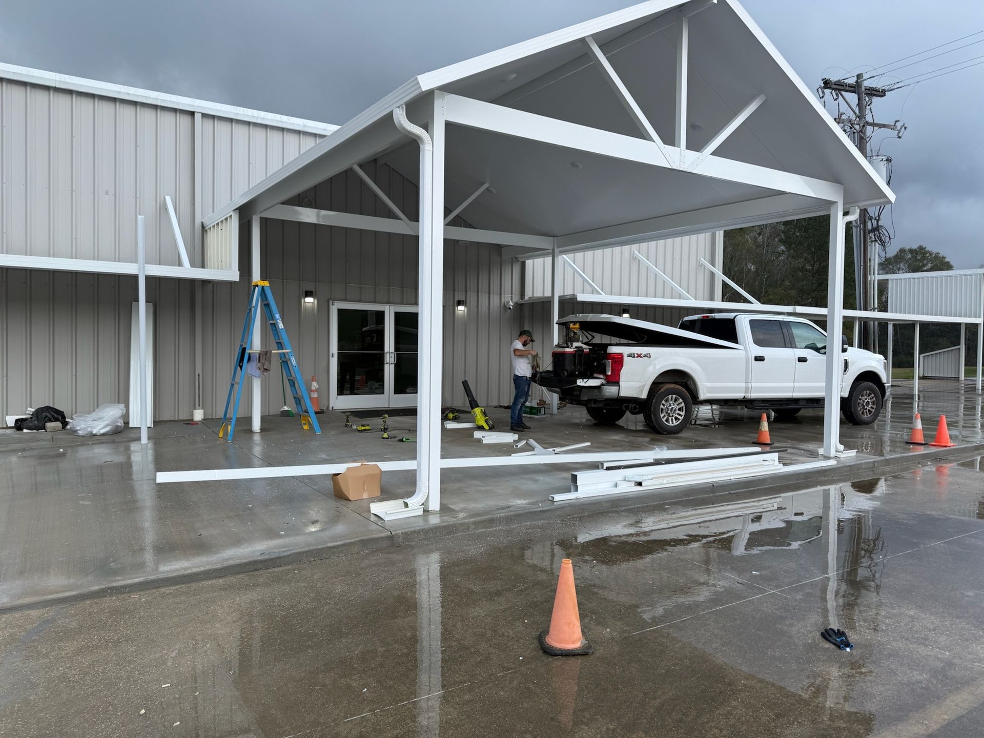 A white canopy being constructed outside a building. Workers and a truck are present. The ground is wet.