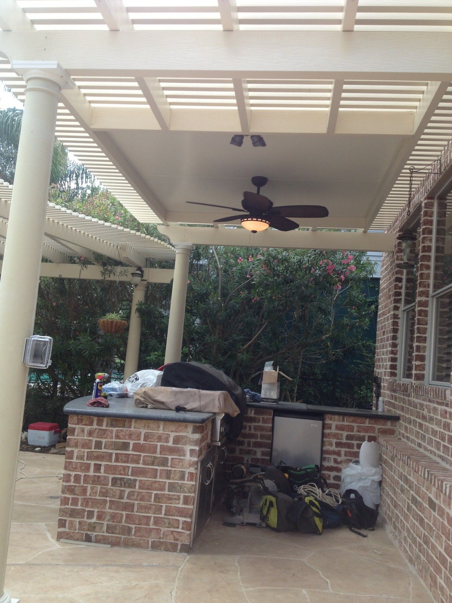 Outdoor kitchen under a pergola with brick facade. Includes a built-in grill, cabinets, and a ceiling fan.