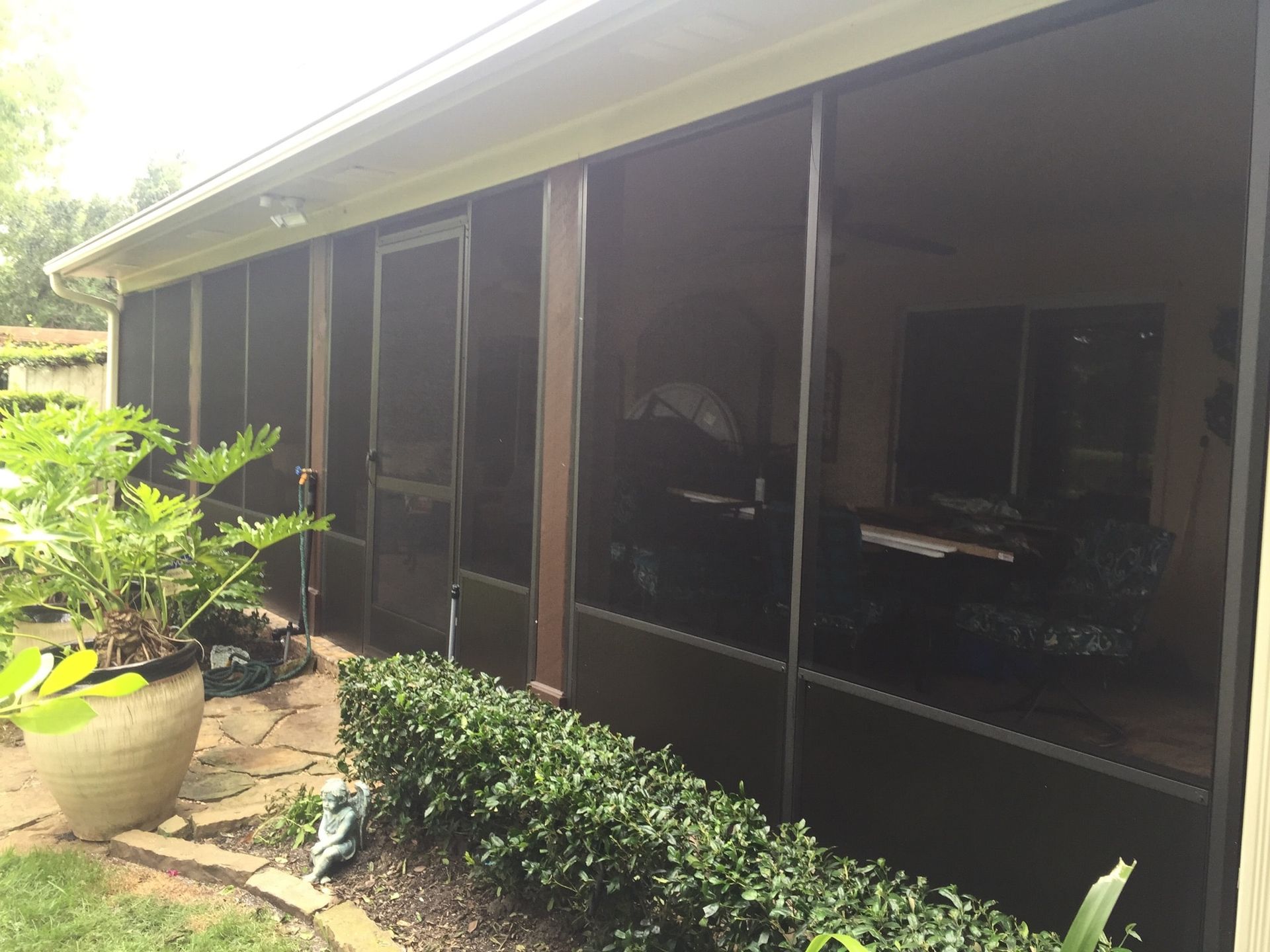 Screened-in porch with brown frames, featuring a potted plant, green hedges, and an interior with furniture.