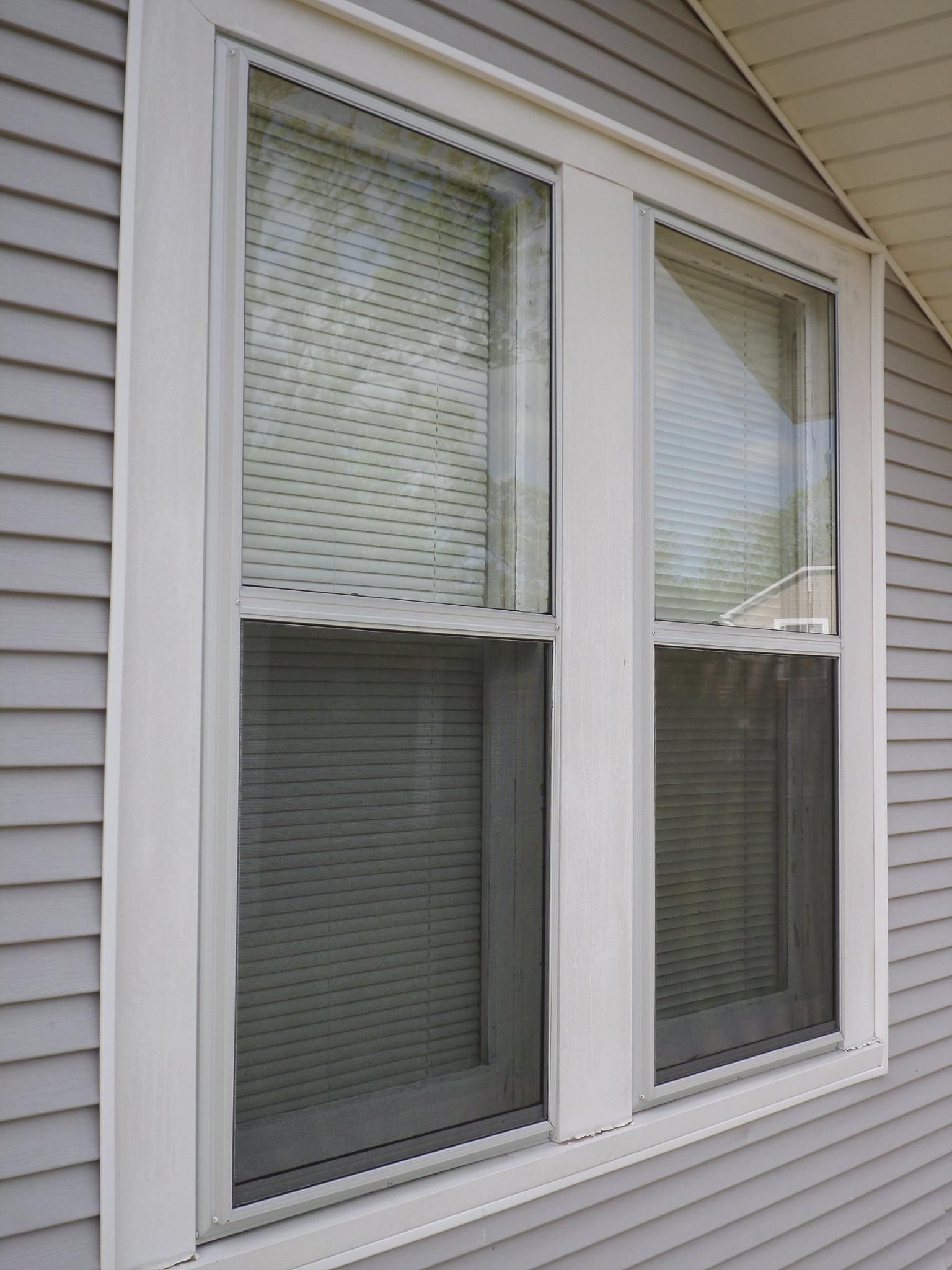 Two double-hung windows with white trim on a gray house. Blinds are visible in both panes.
