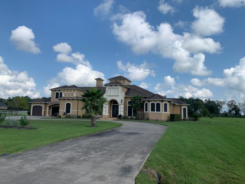 Large beige house with a long driveway on a green lawn under a blue sky with puffy clouds.