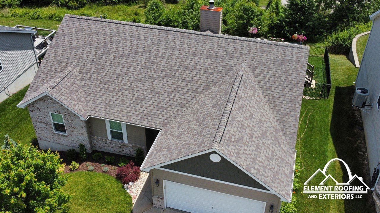 Aerial view of a residential house with a newly installed gray shingled roof and a chimney, surrounded by green lawns.