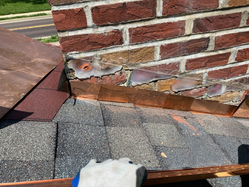 A close-up of a damaged brick chimney base on a shingled roof, showing deteriorated mortar and exposed flashing.