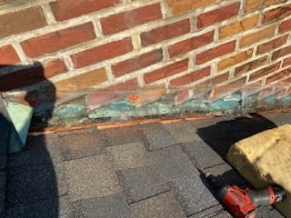 A power drill sits on shingled roofing next to a brick wall with exposed flashing and insulation.