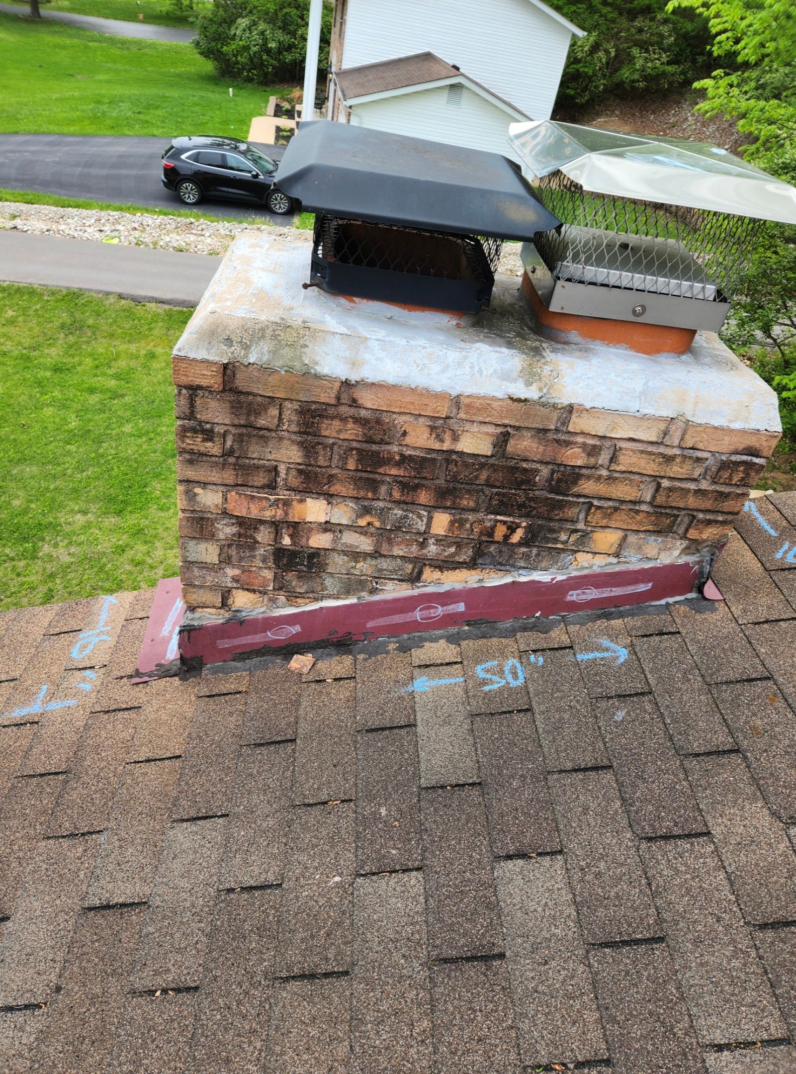 A brick chimney on a shingled roof with a black metal cap, red flashing at the base, and a white shed in the background.