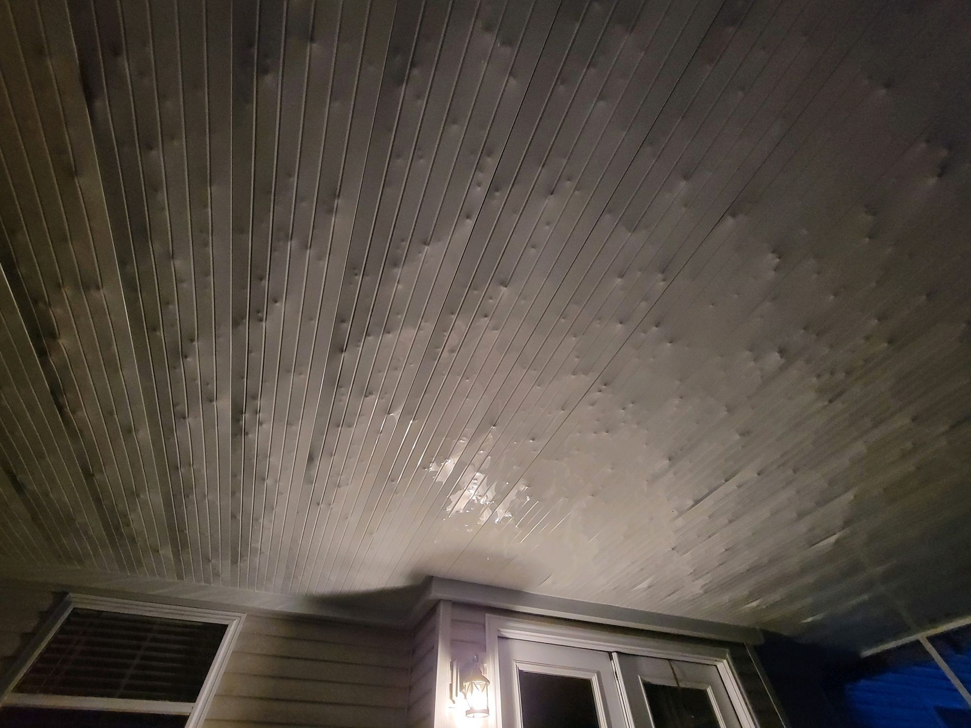 A view looking up at a dark wood-paneled porch ceiling at night, with patches of peeling paint and a light fixture below.