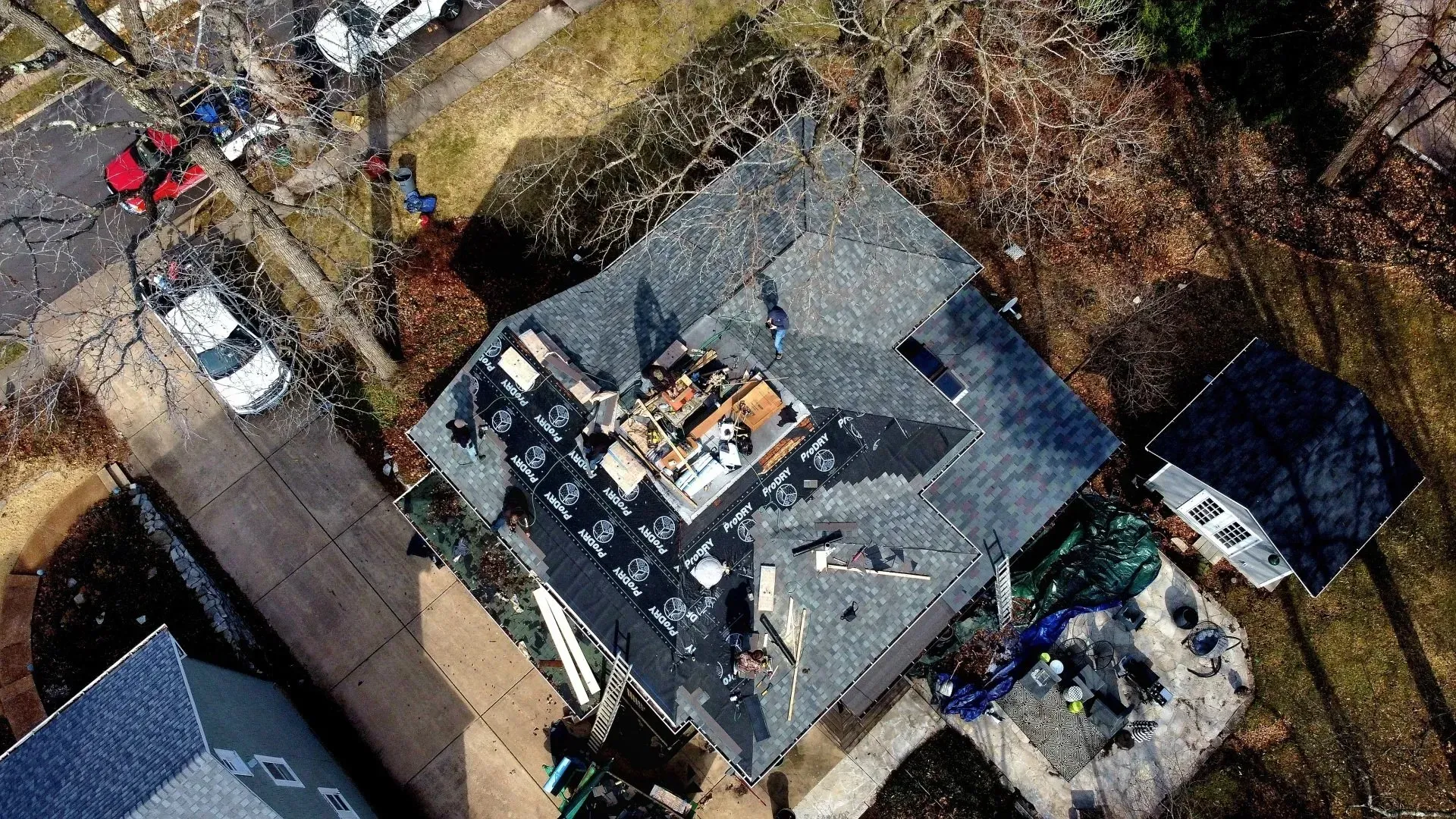 Aerial view of a residential house roof undergoing repairs, showing dark shingles partially removed, tools, and materials.