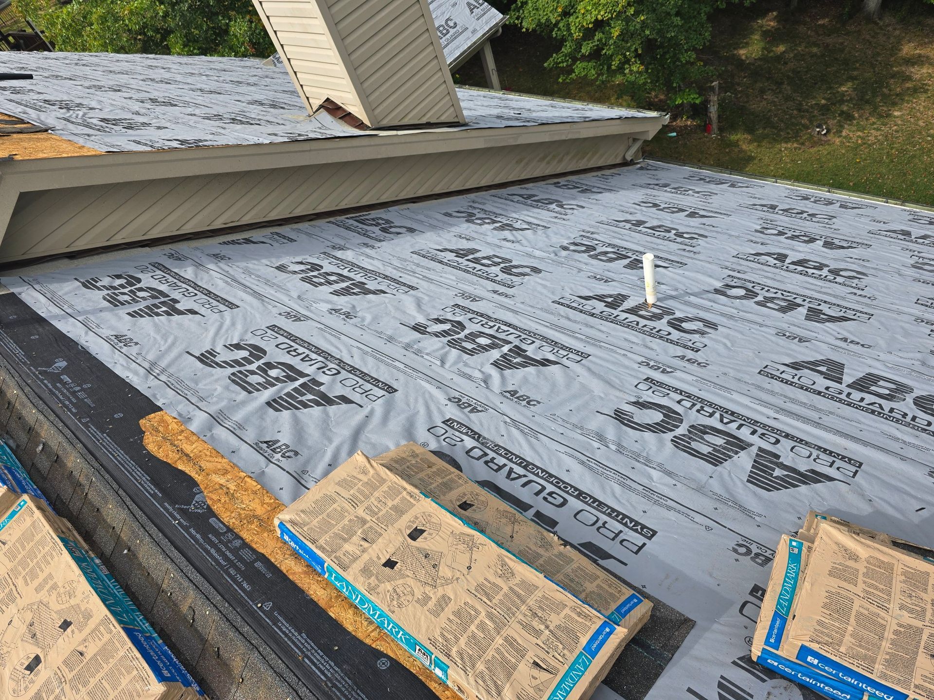 A roof under renovation, covered with grey underlayment, a chimney, and stacks of new roofing shingles.