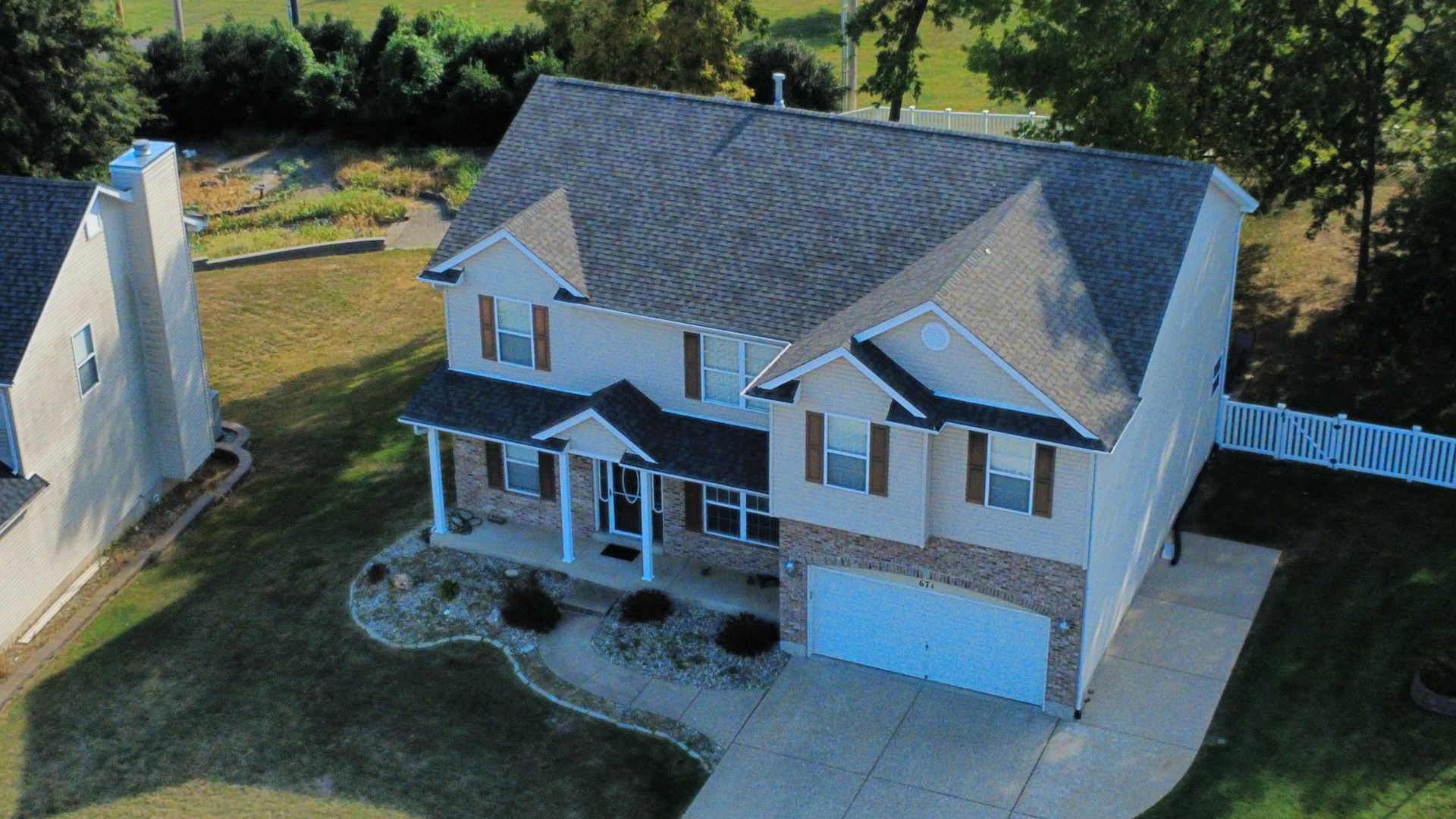 An aerial view of a two-story beige house with brick accents, a gray shingled roof, a front porch, and a two-car garage.