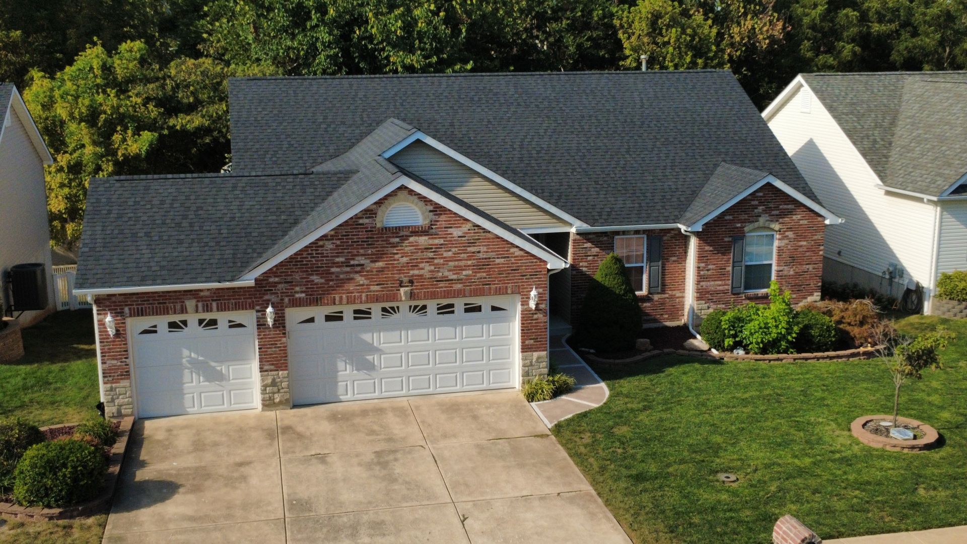 A one-story brick house with a dark shingled roof, a two-car garage, and a green lawn on a sunny day.