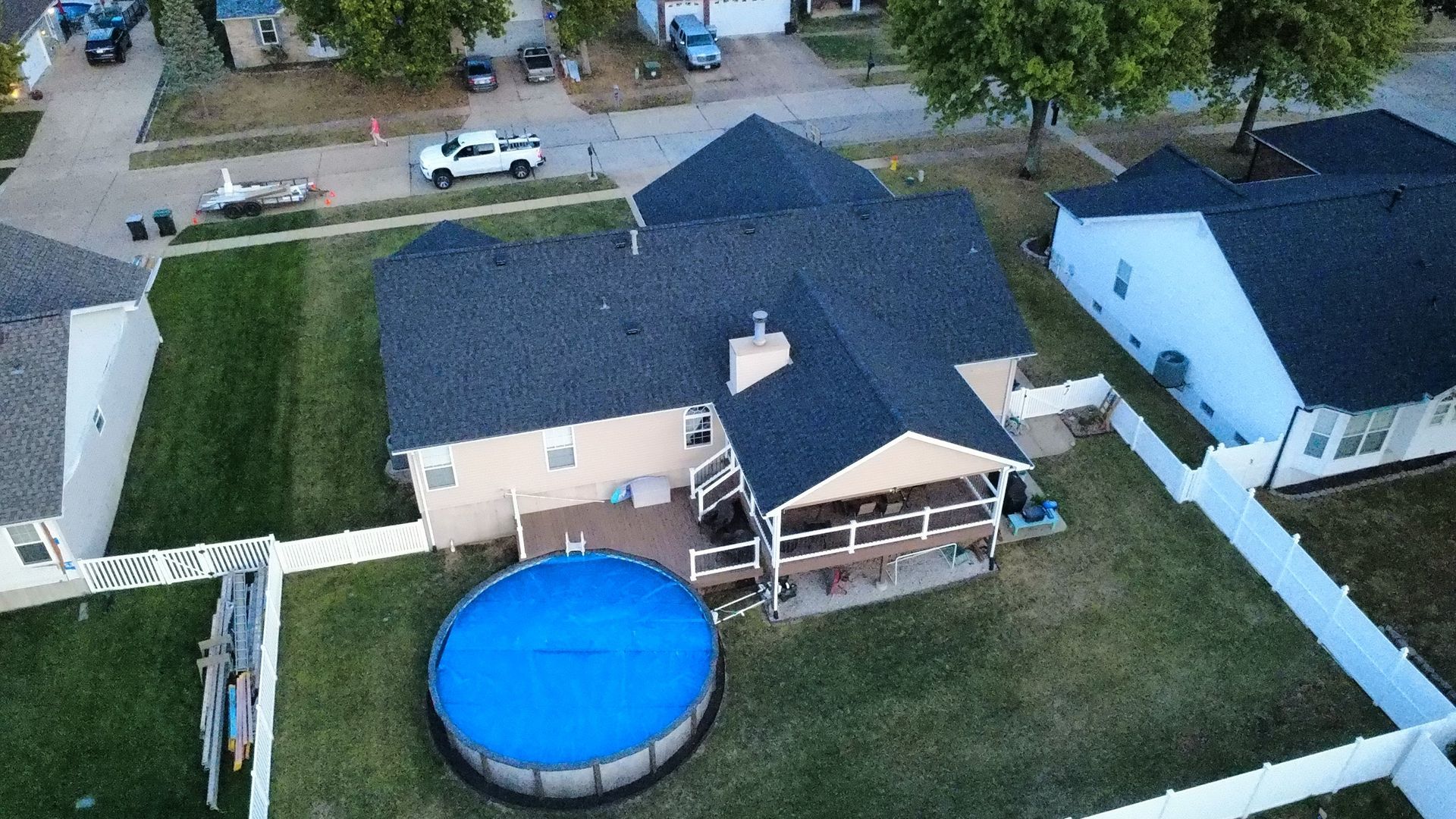 Aerial view of a house with a dark shingled roof, a back deck, and a round above-ground swimming pool in a fenced yard.