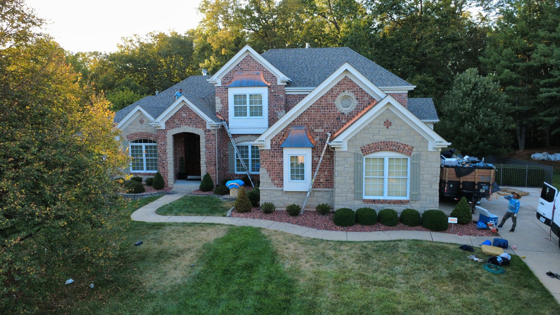 A multi-story brick house with a gray shingled roof, surrounded by trees, with a worker in the driveway during construction.