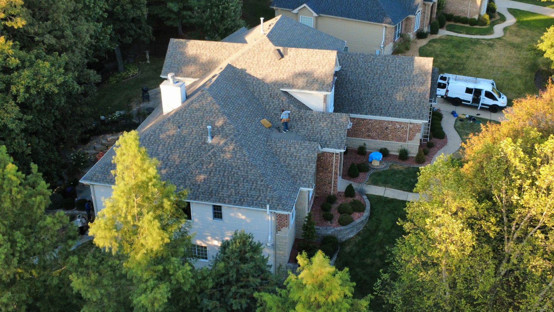 An aerial view of a large residential house with a gray shingled roof and a white van parked in the driveway.