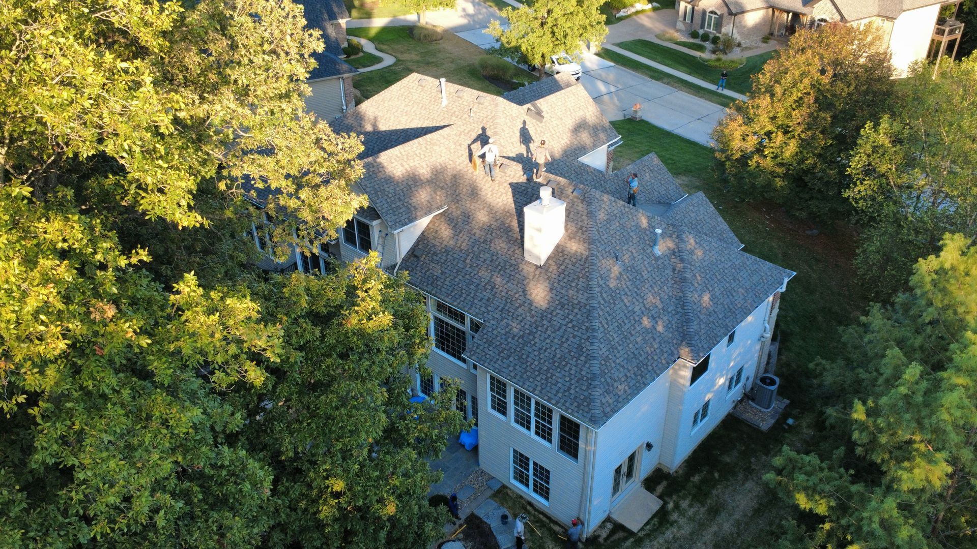 Aerial view of a white, multi-story house with a gray shingled roof, surrounded by green trees and a paved driveway.