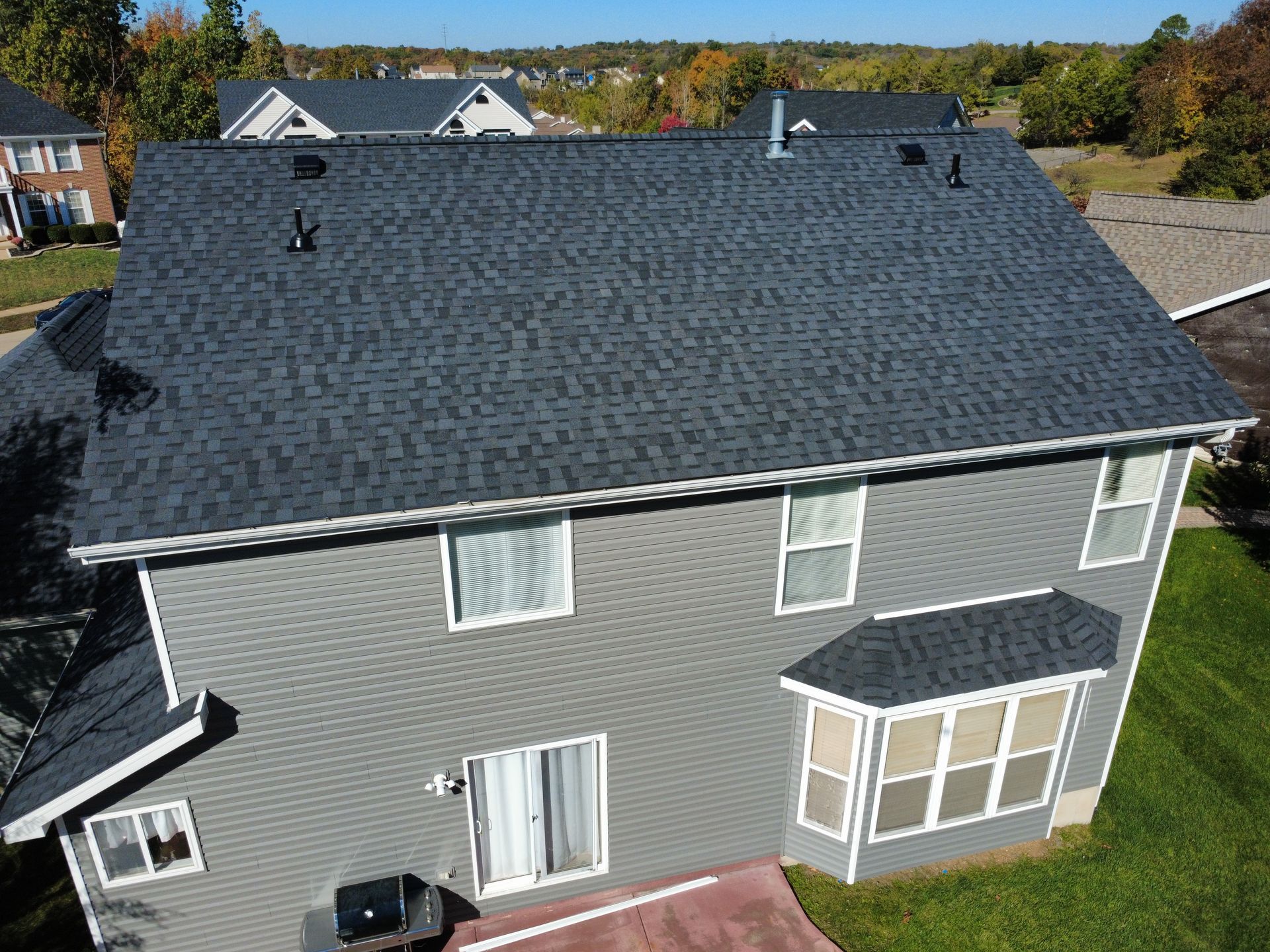 High-angle aerial view of a two-story gray-sided house with a dark shingled roof and a patio, surrounded by green lawns.