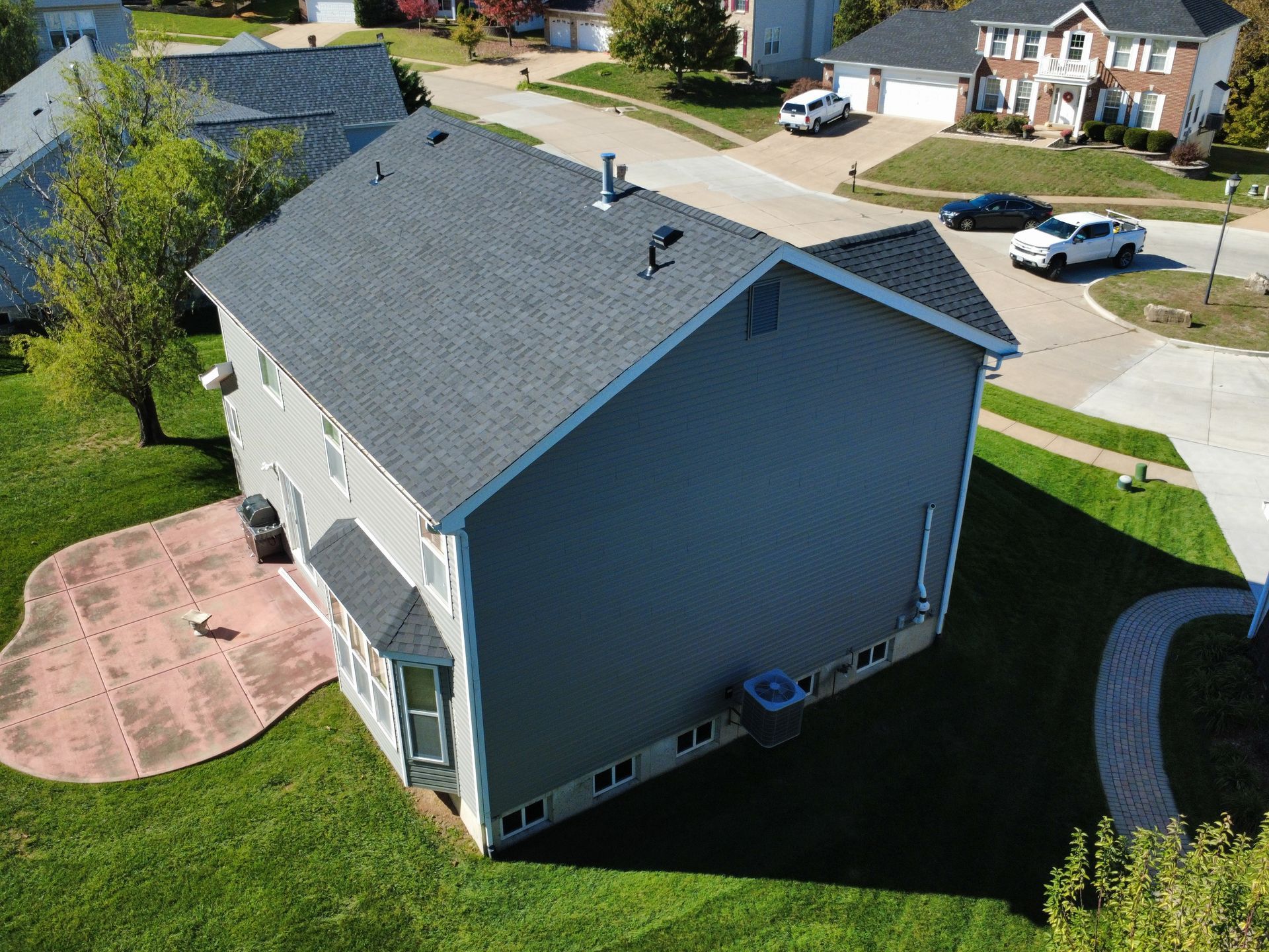 An aerial view of a gray two-story house with a dark shingled roof, a stone patio, and a grassy lawn in a suburban setting.