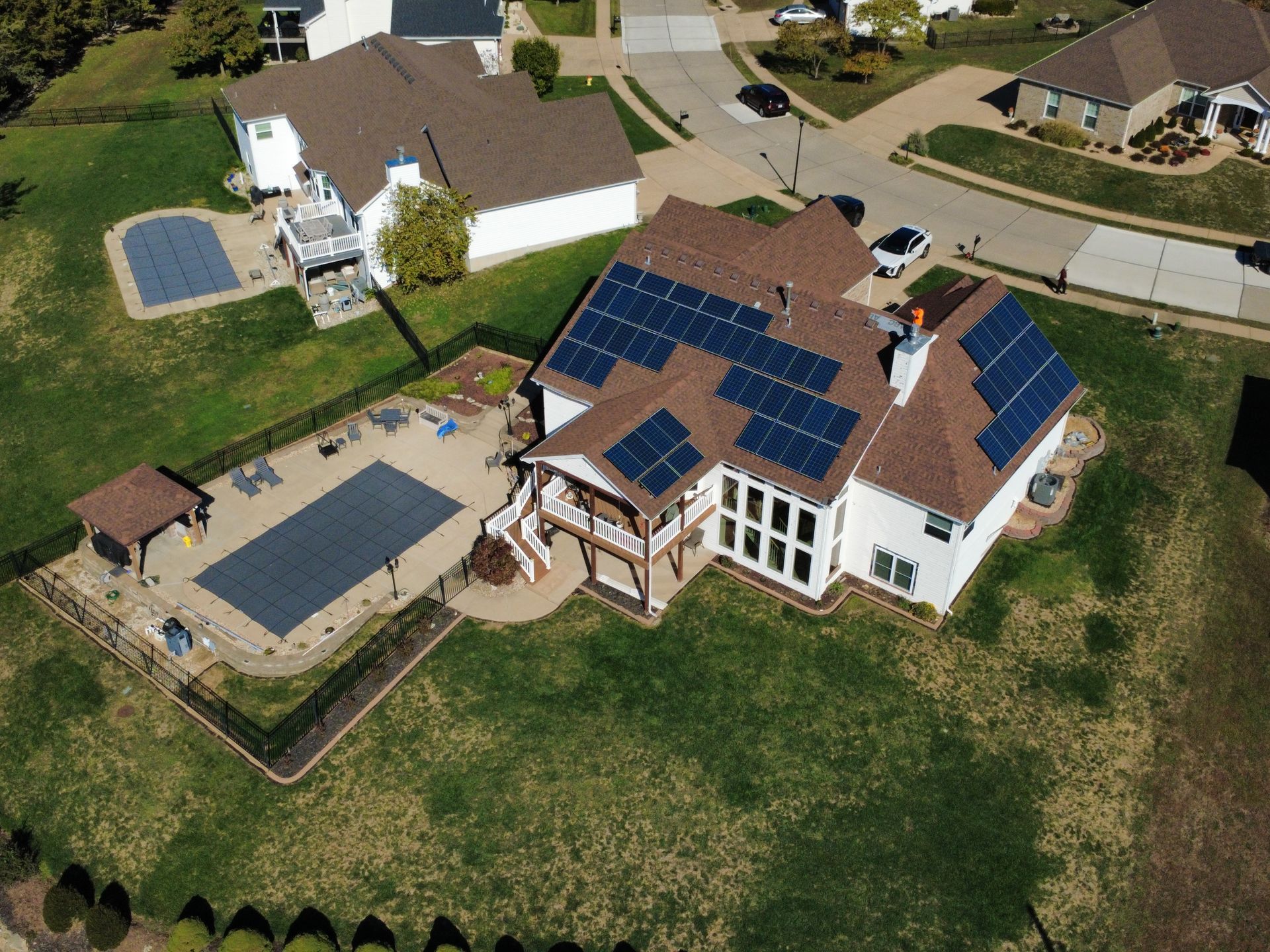 An aerial view of a suburban house featuring solar panels on the roof, a fenced backyard pool, and a gazebo.