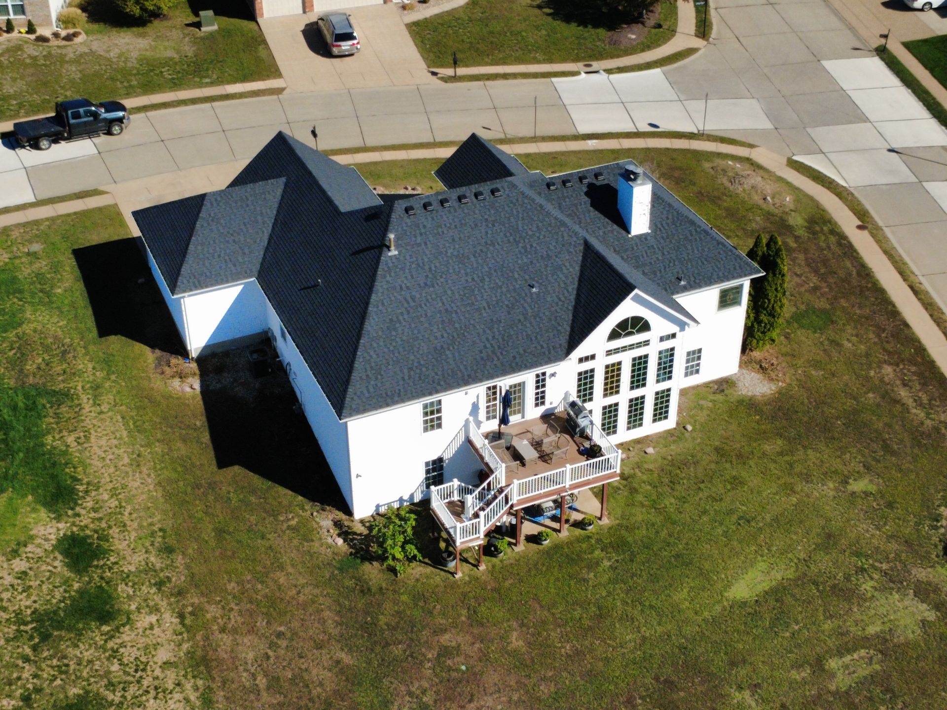 Aerial view of a white house with a dark roof, a wooden deck, and a fenced yard in a suburban neighborhood.