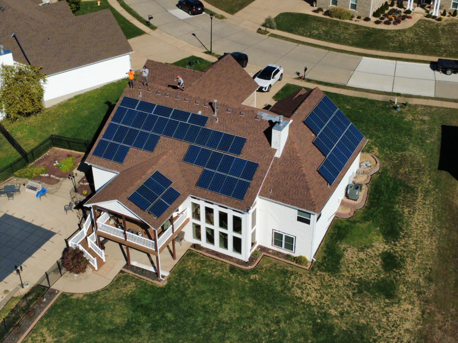 Aerial view of a white house with solar panels installed on its brown shingled roof, surrounded by a lawn and driveway.