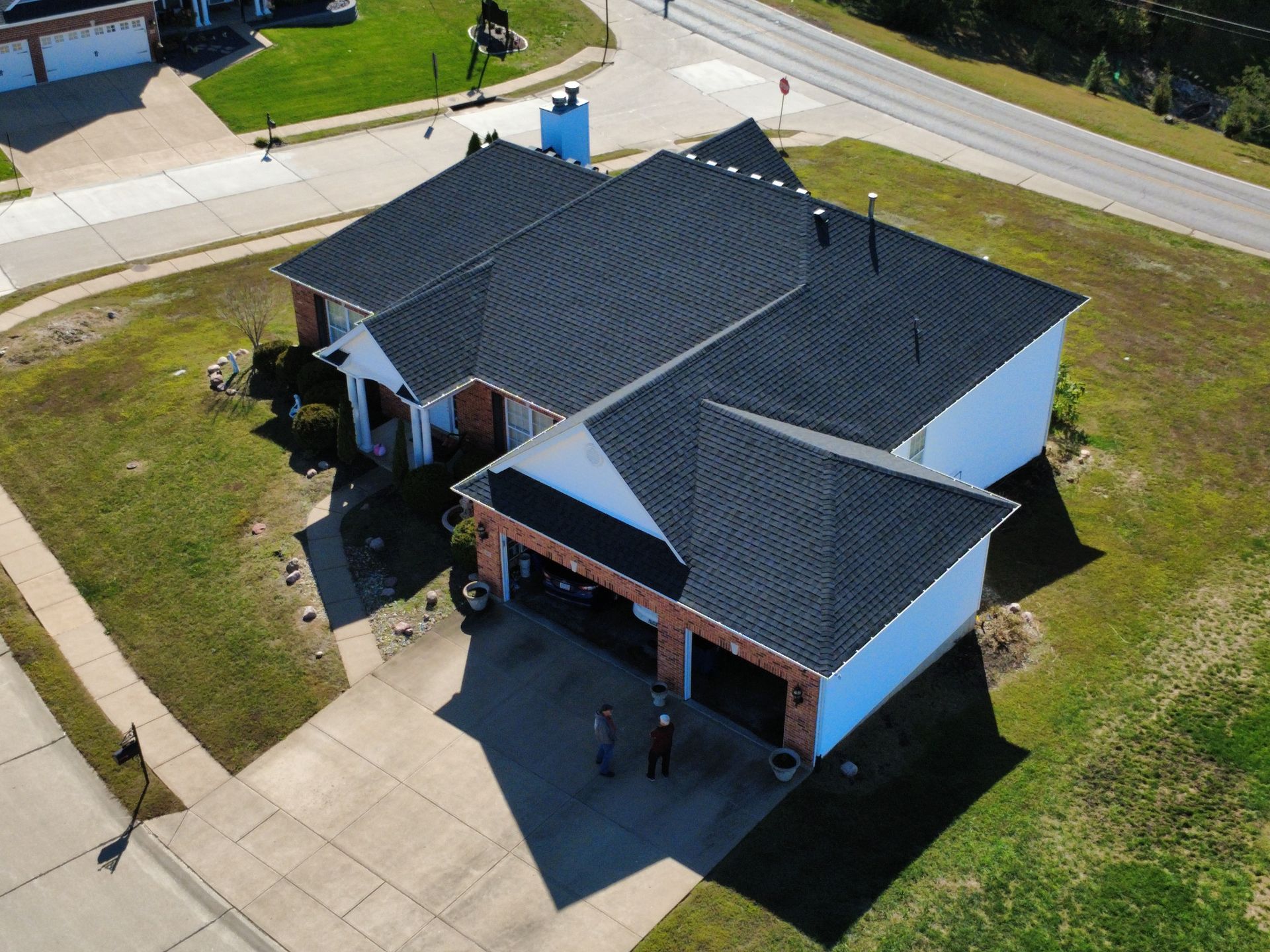 An aerial view of a brick house with a dark shingled roof, featuring a two-car garage and a driveway in a suburban area.